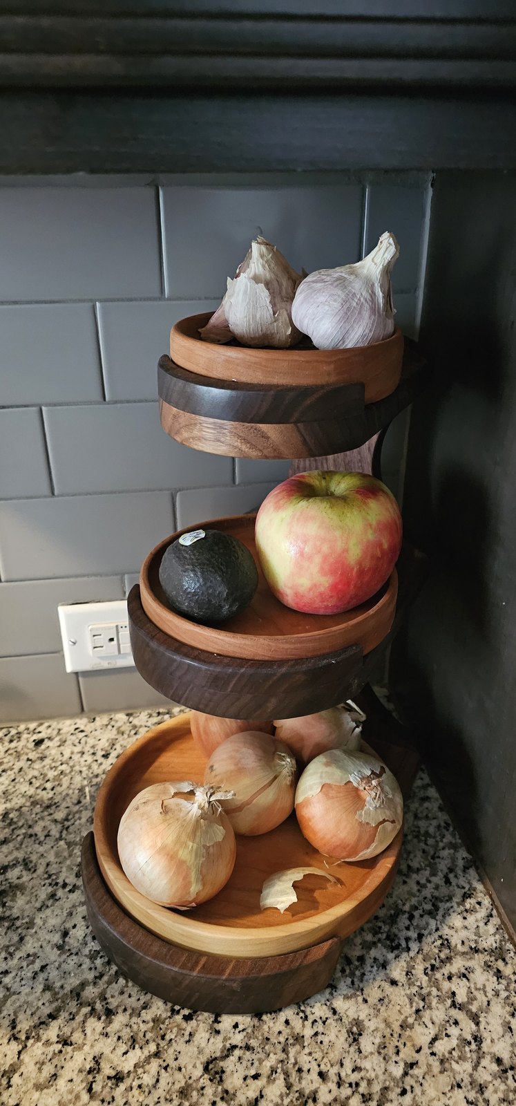 Three-tier wooden fruit stand with light wood bowls on dark wood rings, on a granite counter
